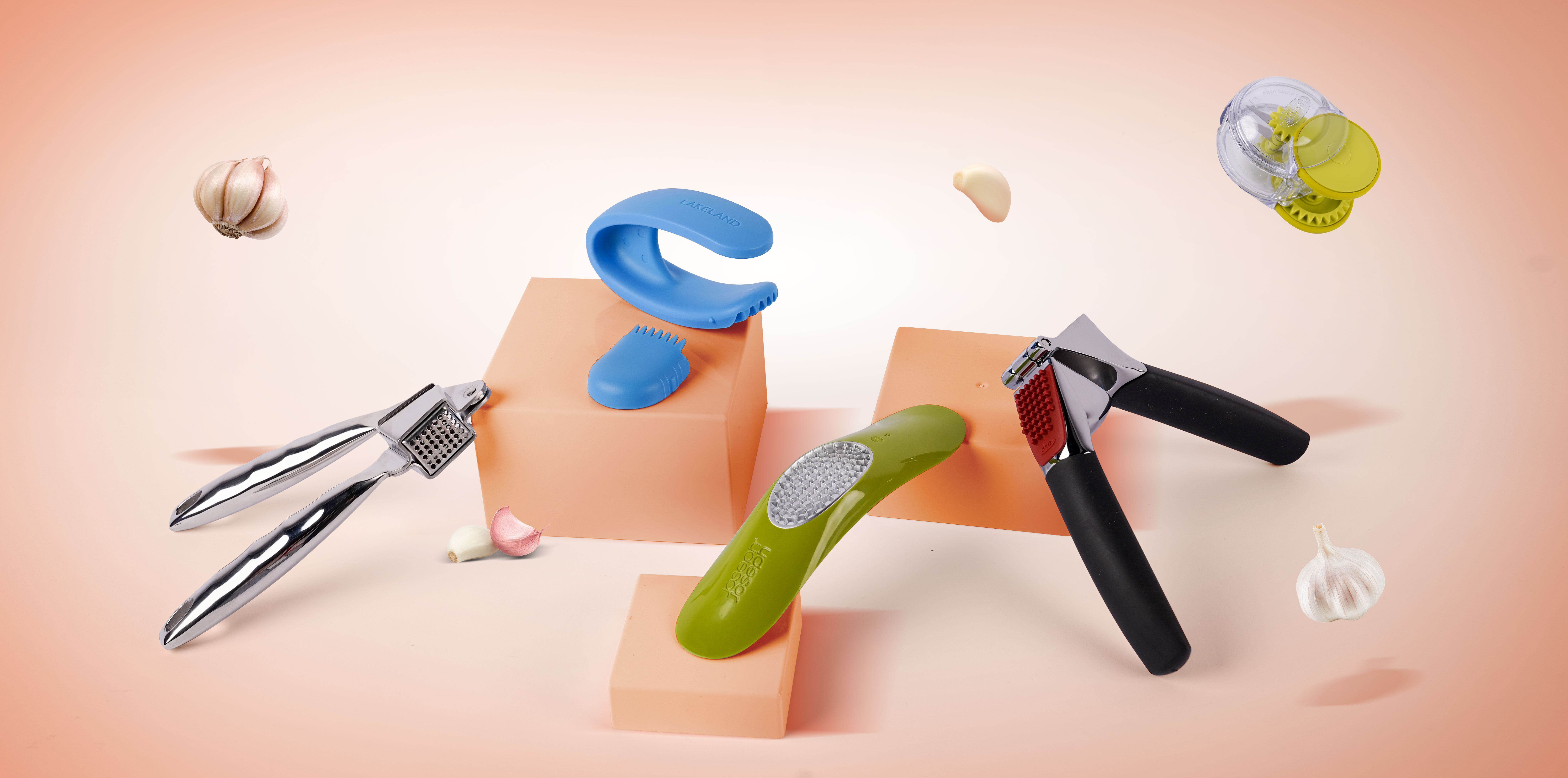 Kitchen tools including garlic presses, rockers and mincers on pink background with floating garlic cloves.