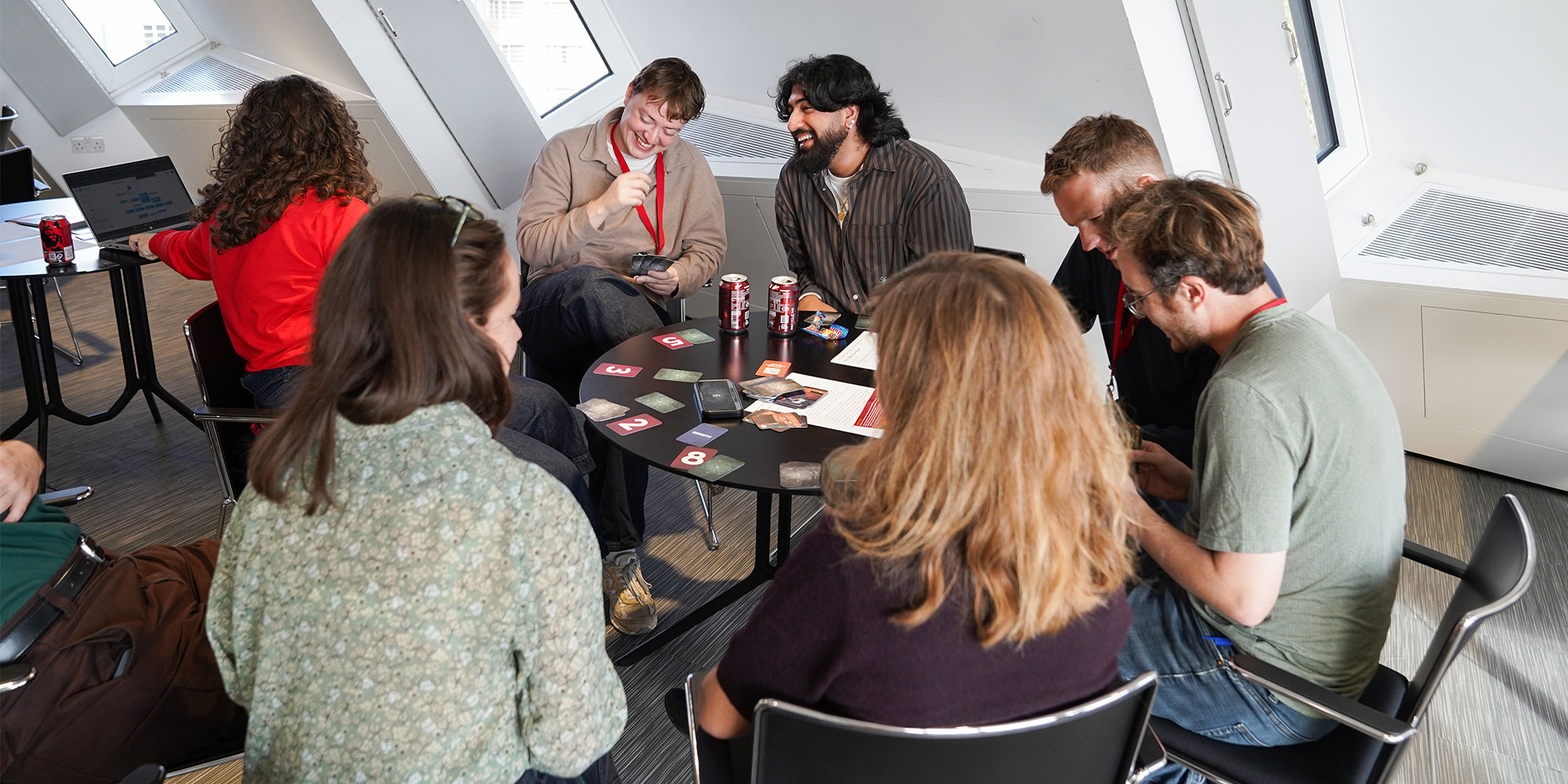 Group of people sitting around a table with cards and cans, engaged in a card game