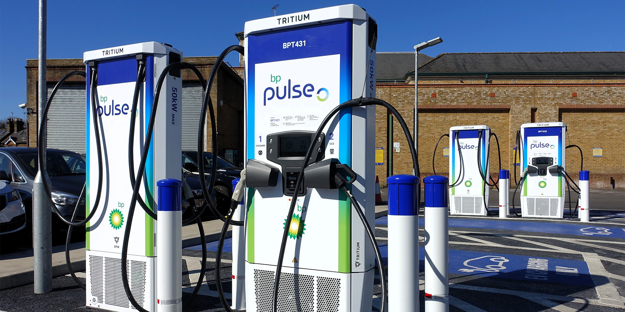 Three electric vehicle charging stations with "bp pulse" branding, positioned in a parking lot with parked cars and a brick building in the background.
