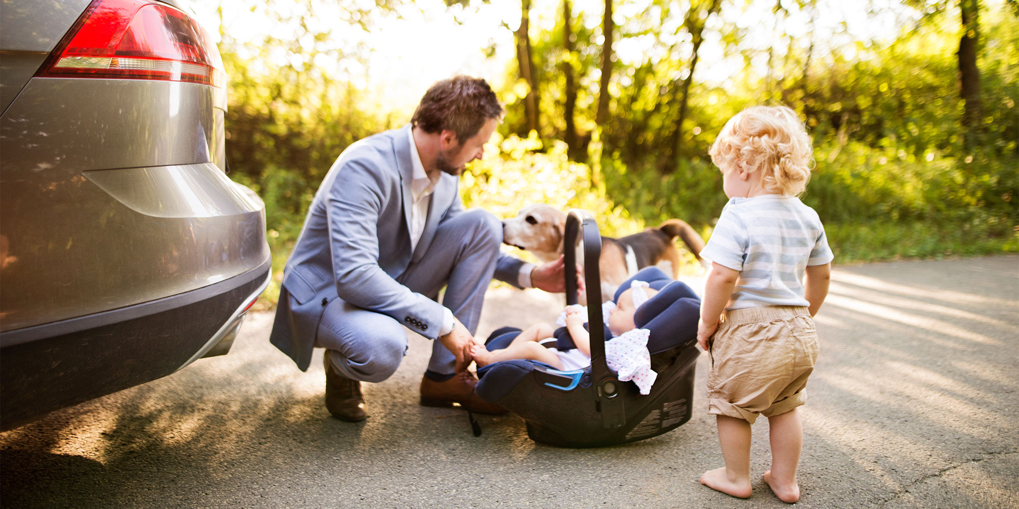 A man in a suit kneels outdoors near a car trunk beside a infant car carrier, with a toddler standing nearby and a dog in the background.