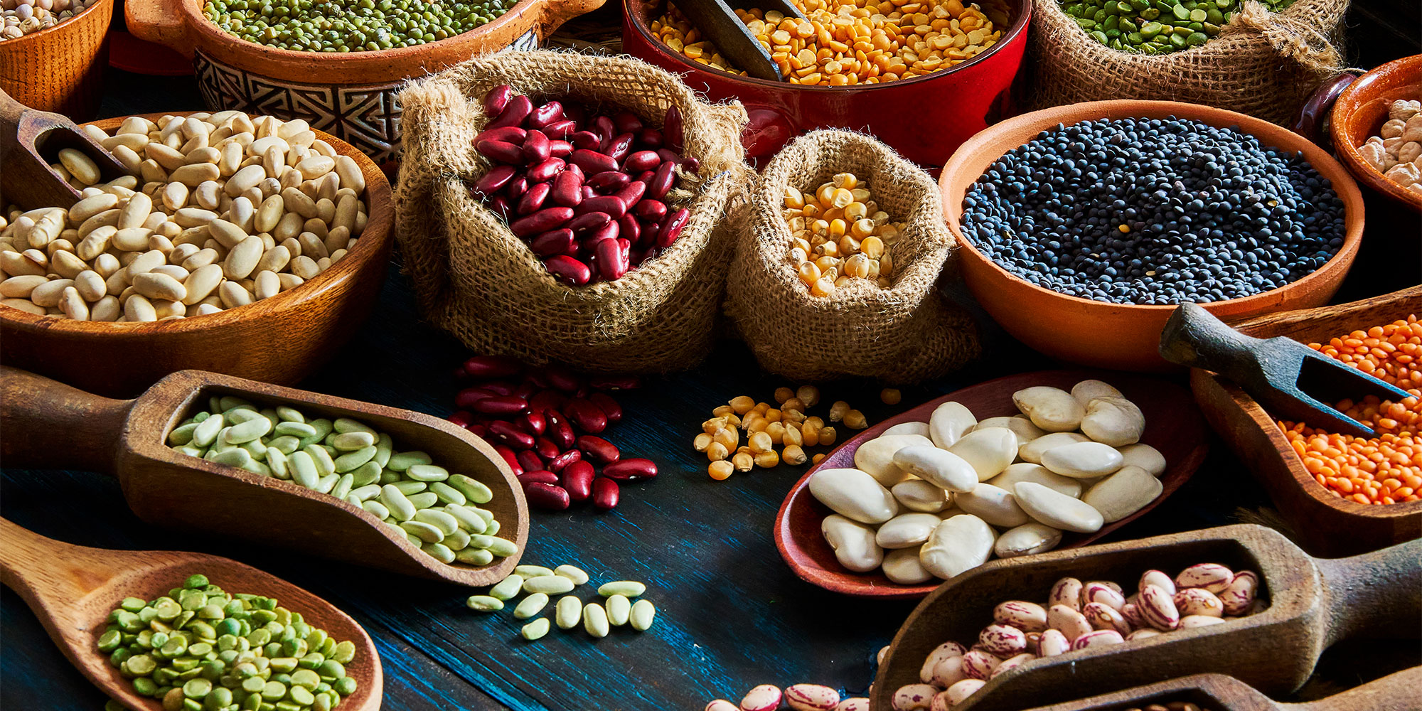 A colourful array of different beans and legumes displayed in various vessels, including bowls, scoops and hessian sacks.