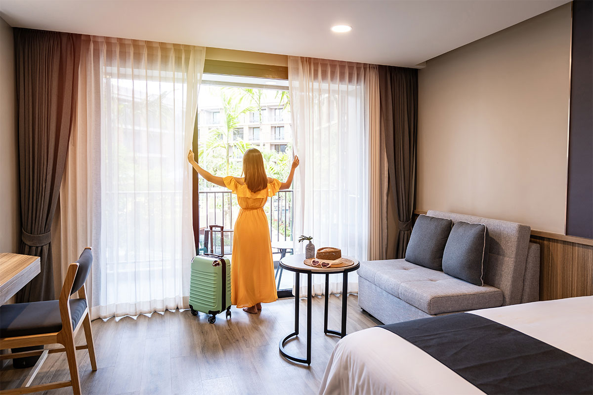 Woman in yellow dress stands with arms outstretched at balcony window in hotel room.