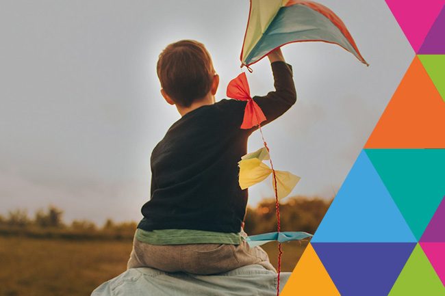 Child flying a colorful kite in an open field, viewed from behind, with a vibrant geometric pattern in the corner.