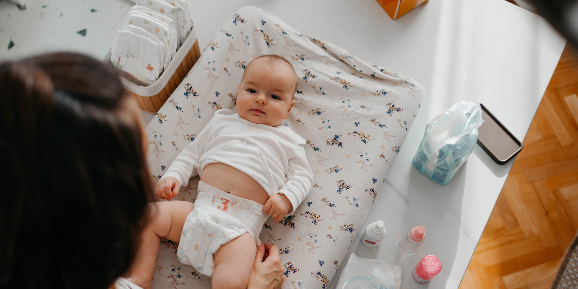 An overhead view of a baby lying on a floral-patterned changing mat. A caregiver's hands are visible adjusting the baby’s nappy. Nearby are nappies, wipes, and baby care bottles.