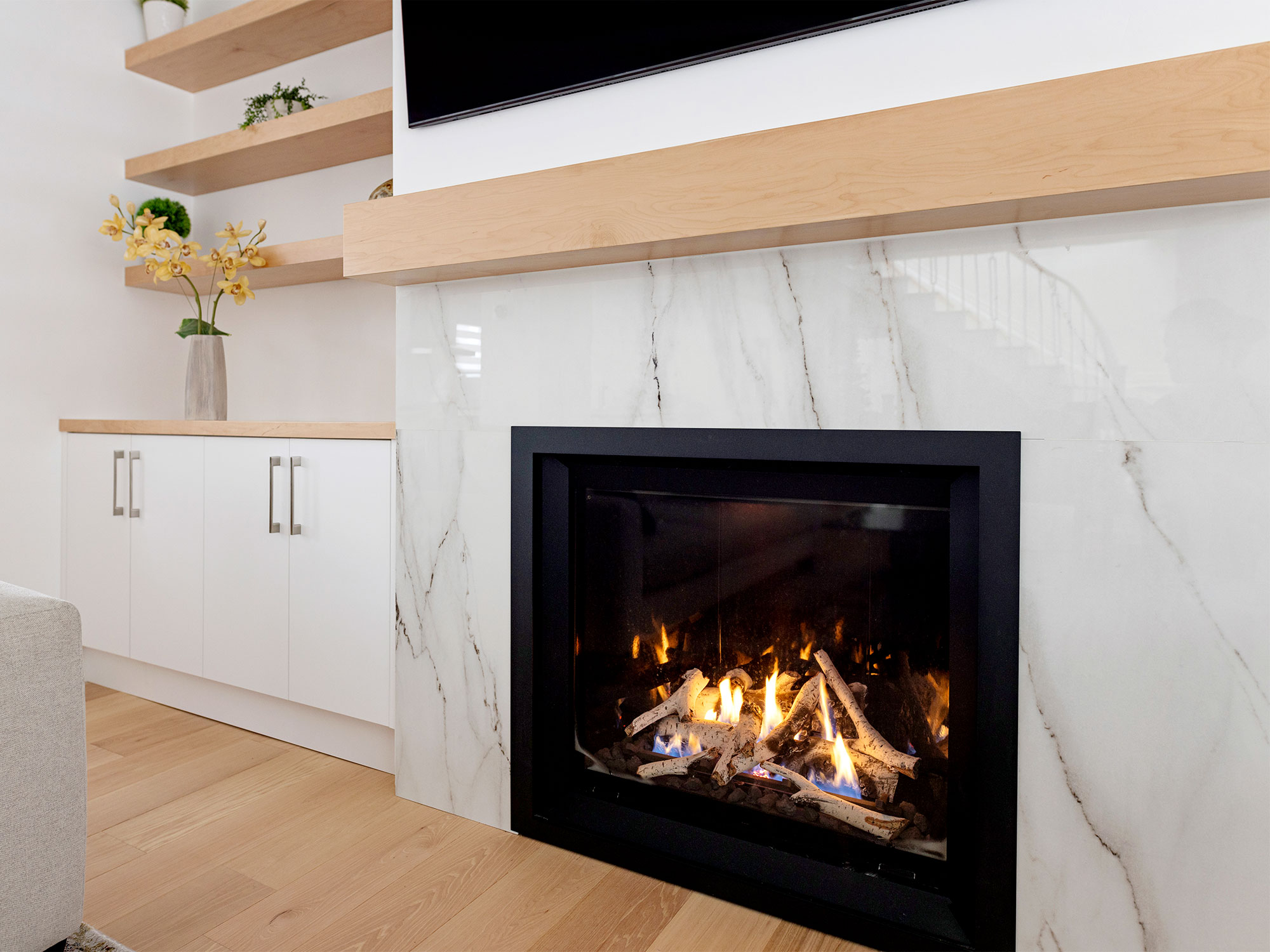Modern living room with white marble fireplace, black frame, and glowing fire. Adjacent to wooden shelves and white cabinets.