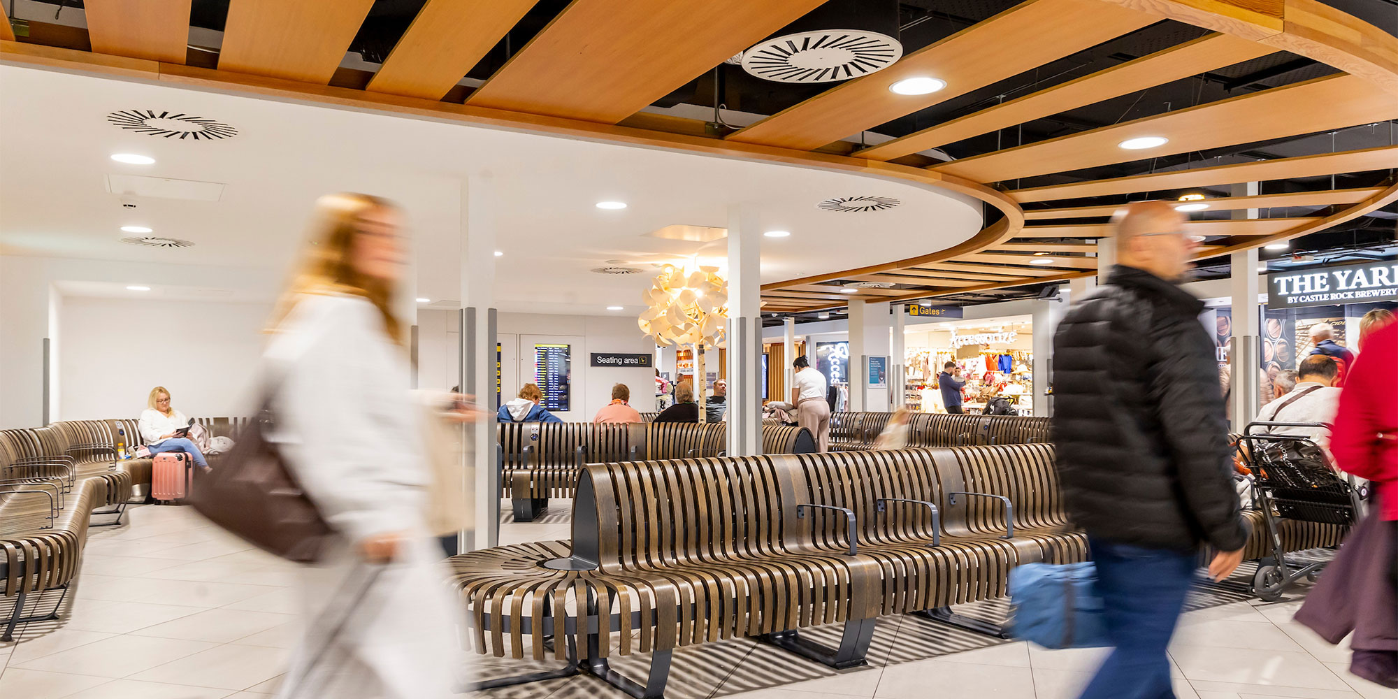 Busy airport lounge with people walking, sitting, and shopping. Modern design with wooden benches and circular ceiling lights.