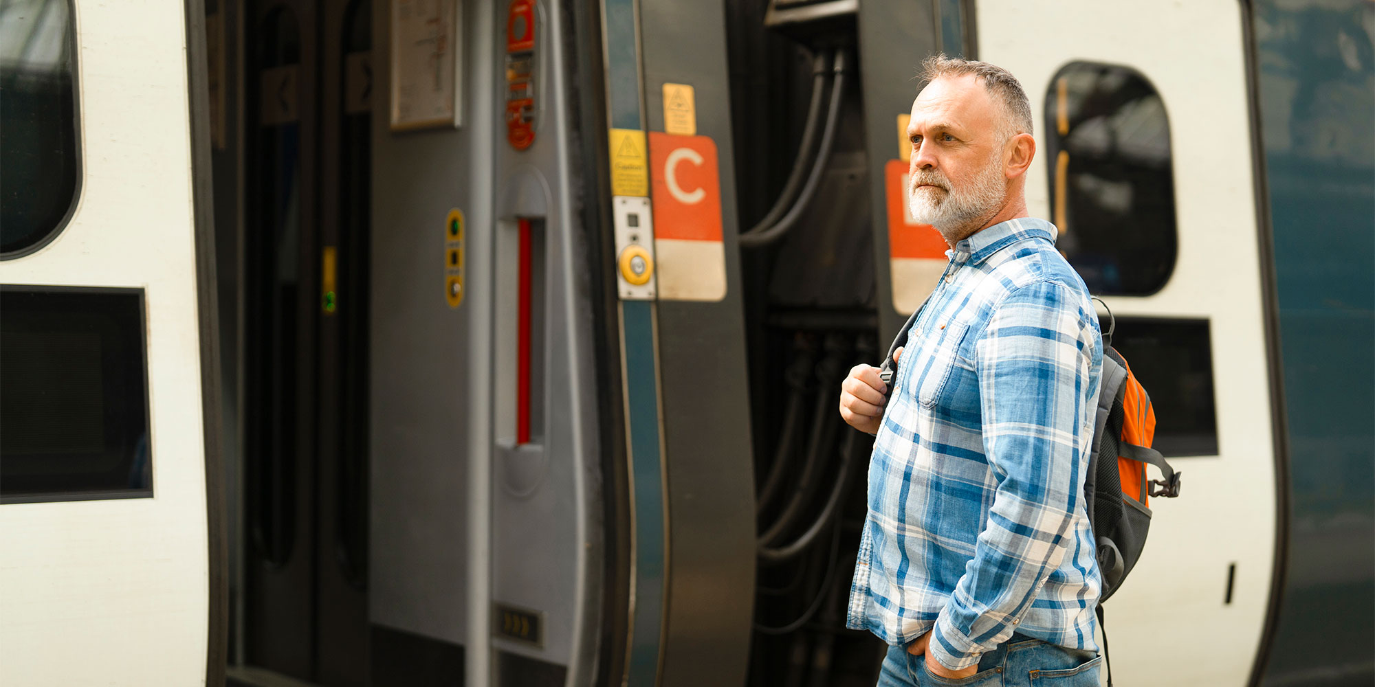 Man with backpack stands by train door, hands in pockets, wearing blue plaid shirt and jeans.