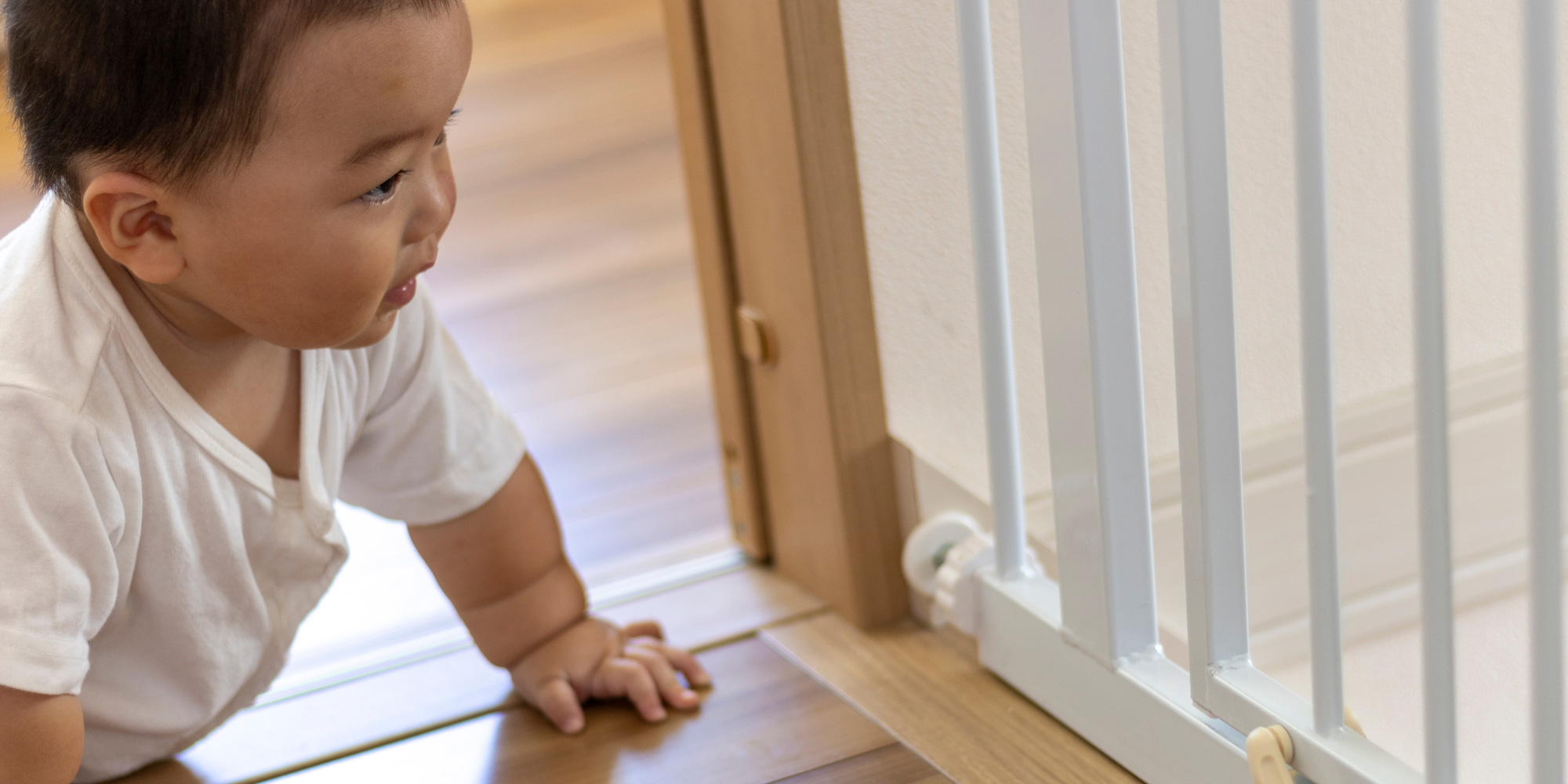 A close-up, side-profile view of a baby crawling on a wooden floor toward a white metal safety gate installed in a doorway.