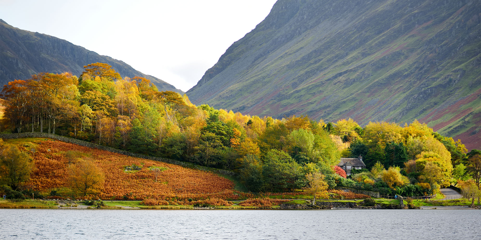 Scenic view of a lake with autumn trees, a stone house, and mountains in the background.