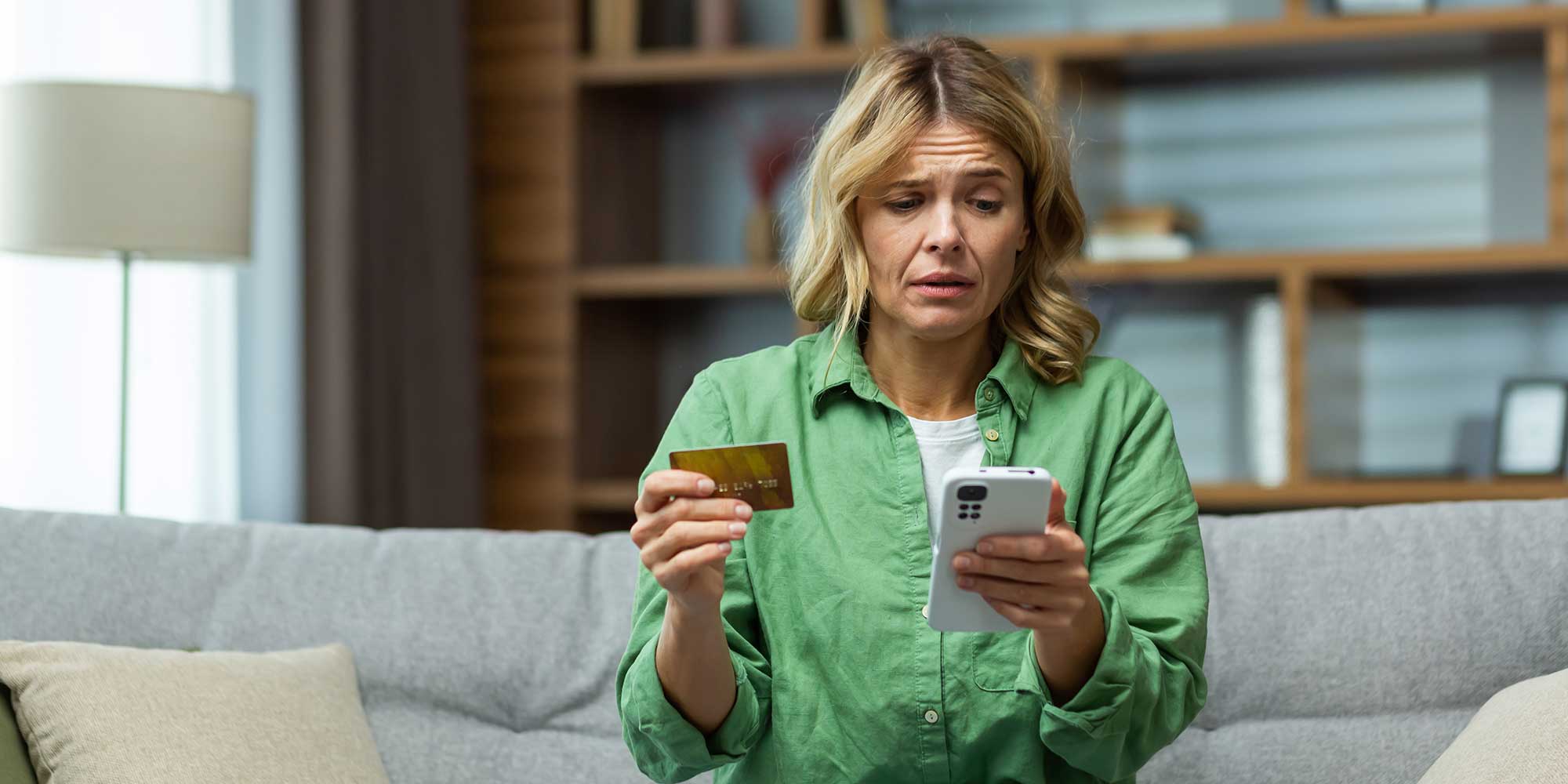 Blonde woman looking stressed holding a debit card and her phone