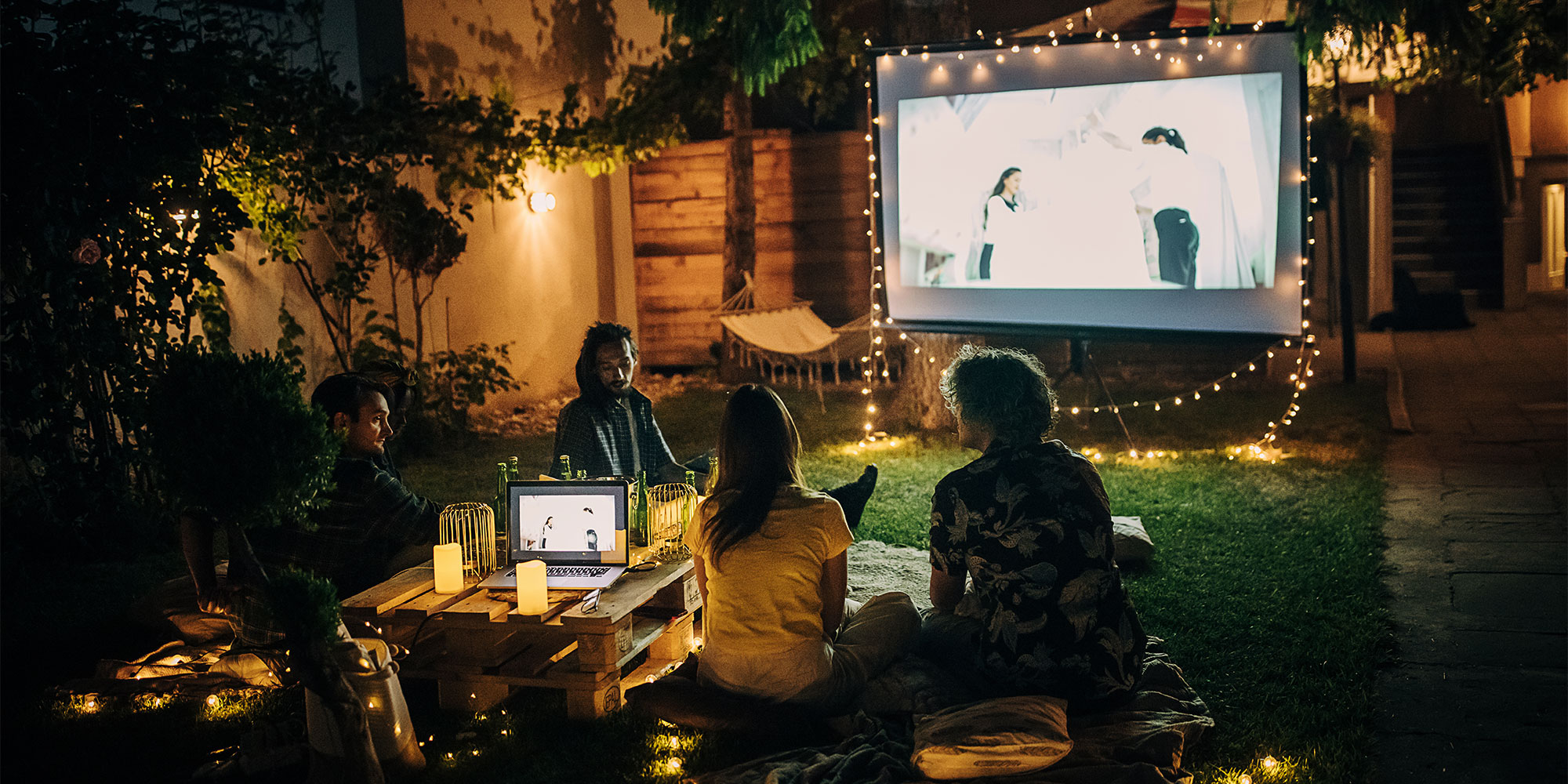 People watching a film on an outdoor projector with string lights and a laptop