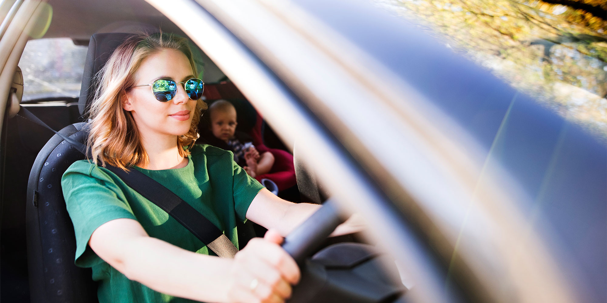 A woman with blonde hair and sunglasses drives a vehicle while a baby sits in a red car seat in the back.