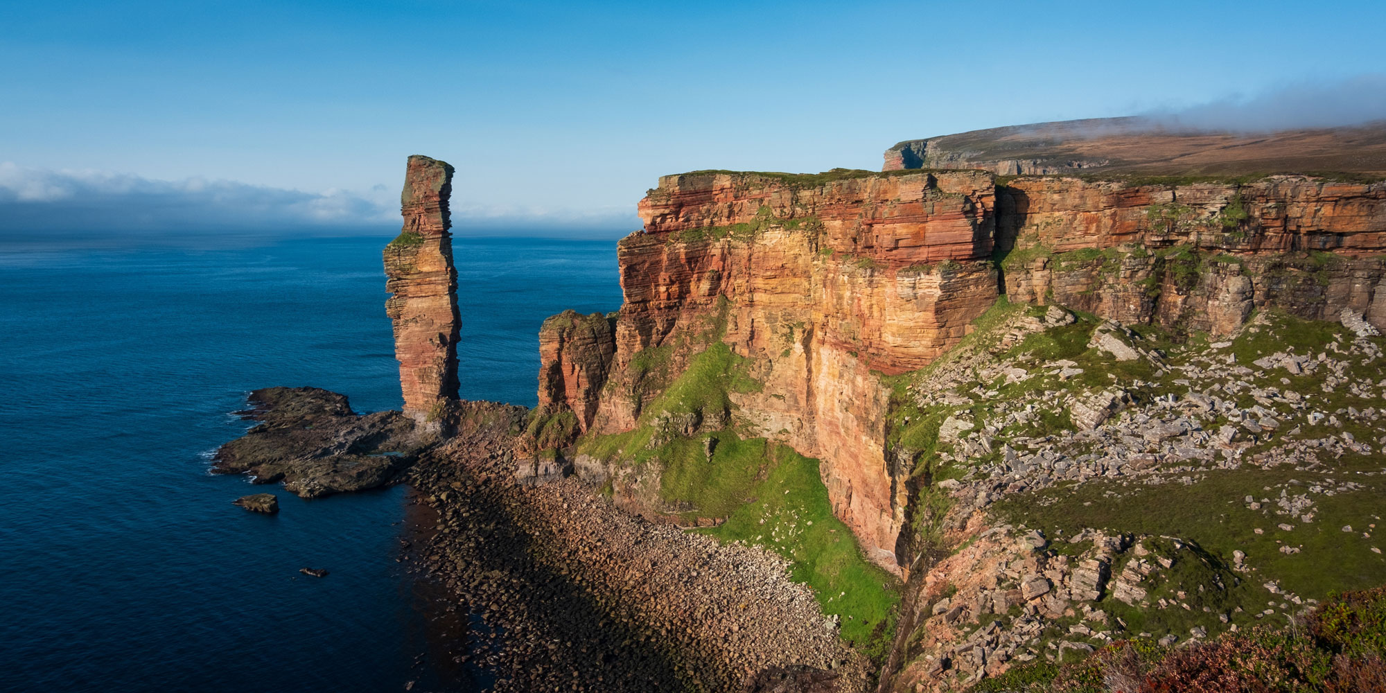Aerial view of rugged cliffs and sea stacks by the ocean under a clear blue sky on the island of Orkney.