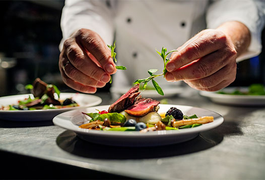 Chef in white uniform garnishing a plate of meat, vegetables, and berries with herbs.