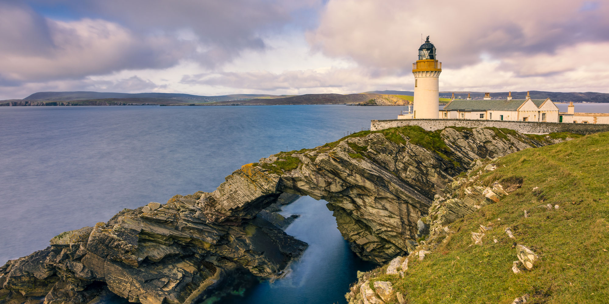 Lighthouse on rocky cliff with natural arch over water, surrounded by hills and cloudy sky.