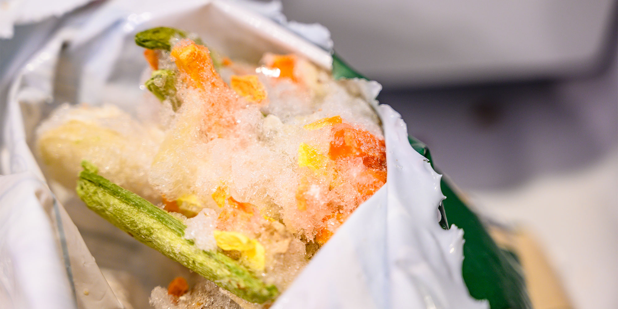 A view inside a plastic bag of mixed frozen vegetables, including carrots and green beans, which are almost entirely obscured by a dense buildup of snowy white ice crystals.