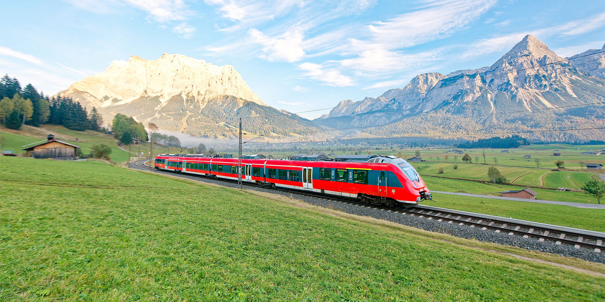 Red passenger train on tracks through a lush green valley with mountains in the background.