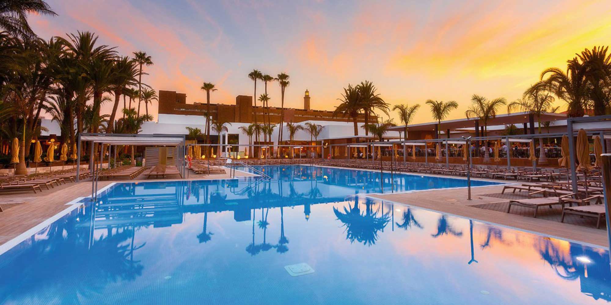 A tranquil pool reflecting palm trees and a sunset sky, surrounded by lounge chairs and a modern resort setting.