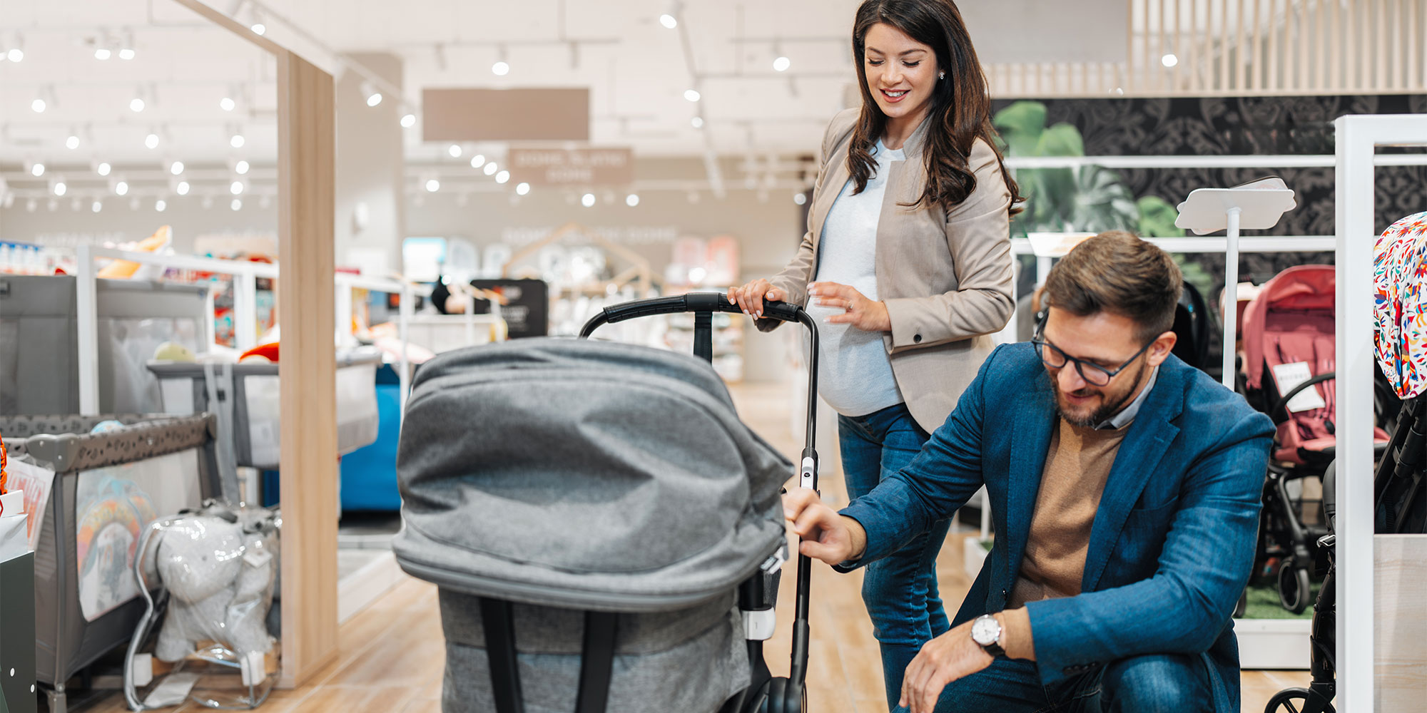 A pregnant woman and a man in a blue blazer look at a gray stroller in a brightly lit baby store. Other nursery furniture and strollers are visible in the background.