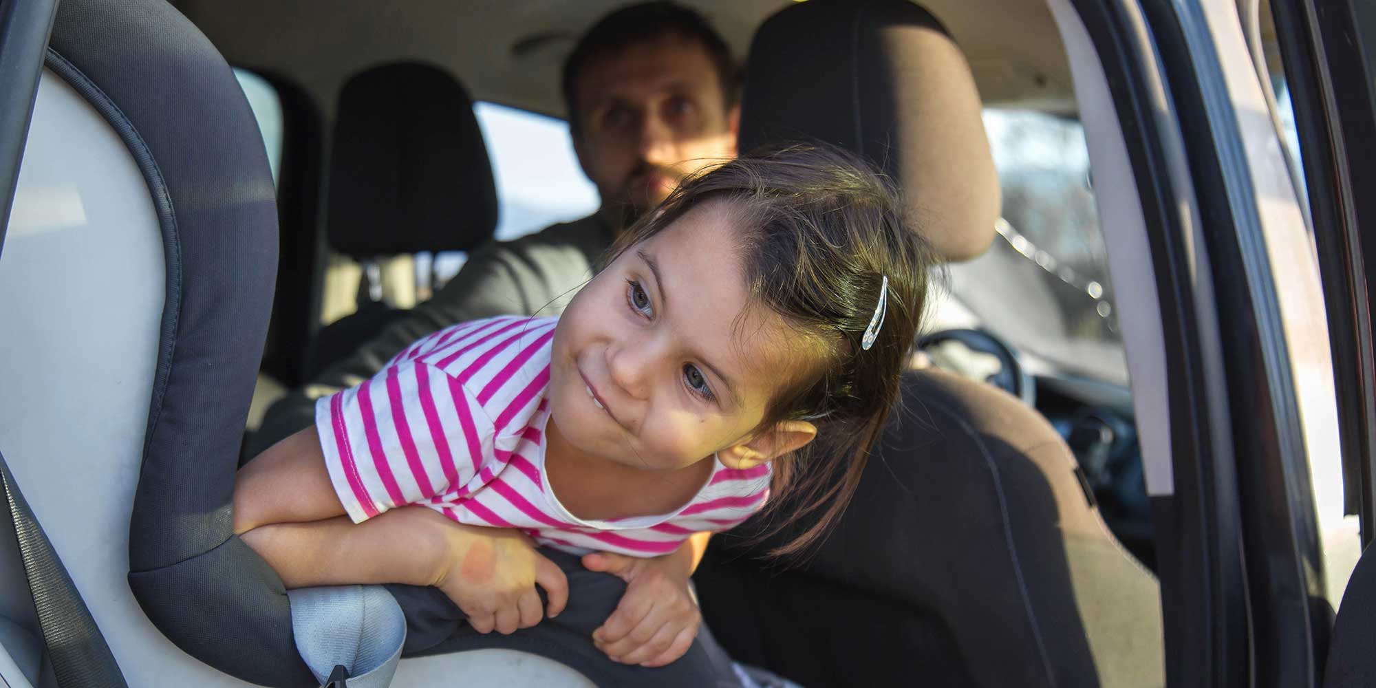 A young girl with dark hair and a pink striped shirt leaning out over her car seat, smiling, with a man visible in the driver's seat background.