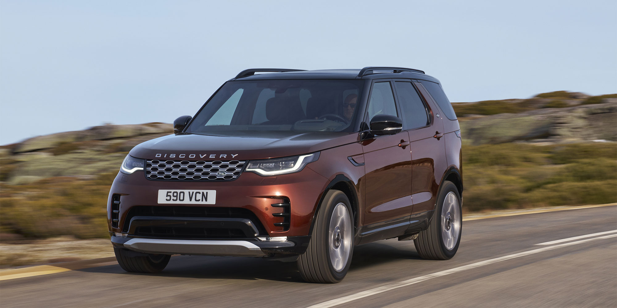 Maroon Land Rover Discovery SUV driving on a road with rocky hills in the background.
