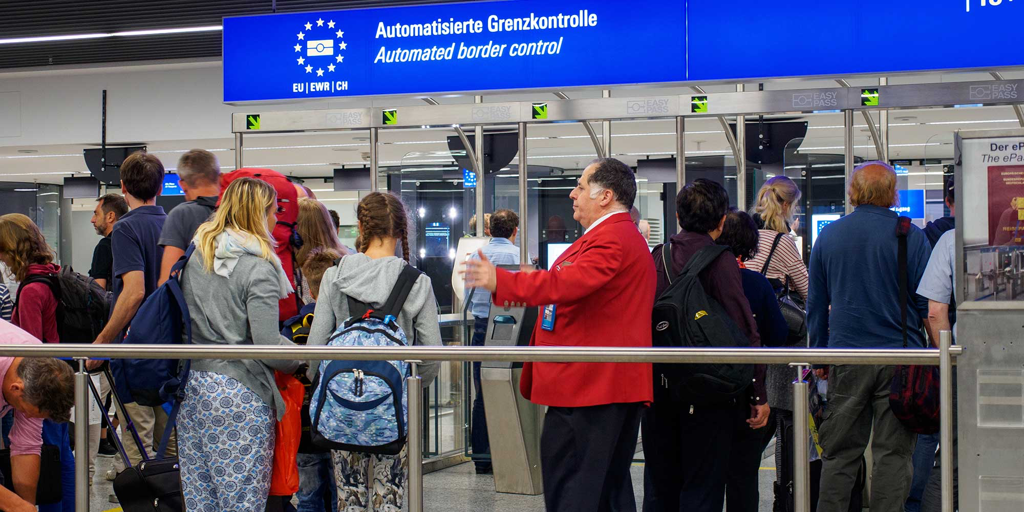 Group of people at an airport border control area, with a man in a red jacket speaking.