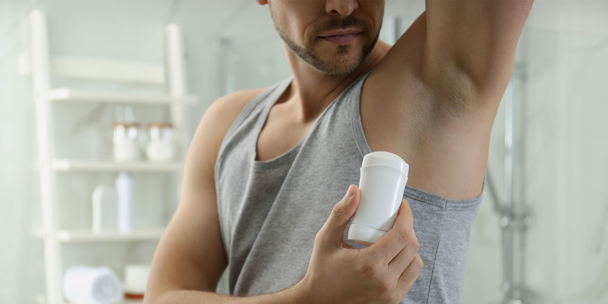 Man in grey tank top applying stick deodorant to armpit