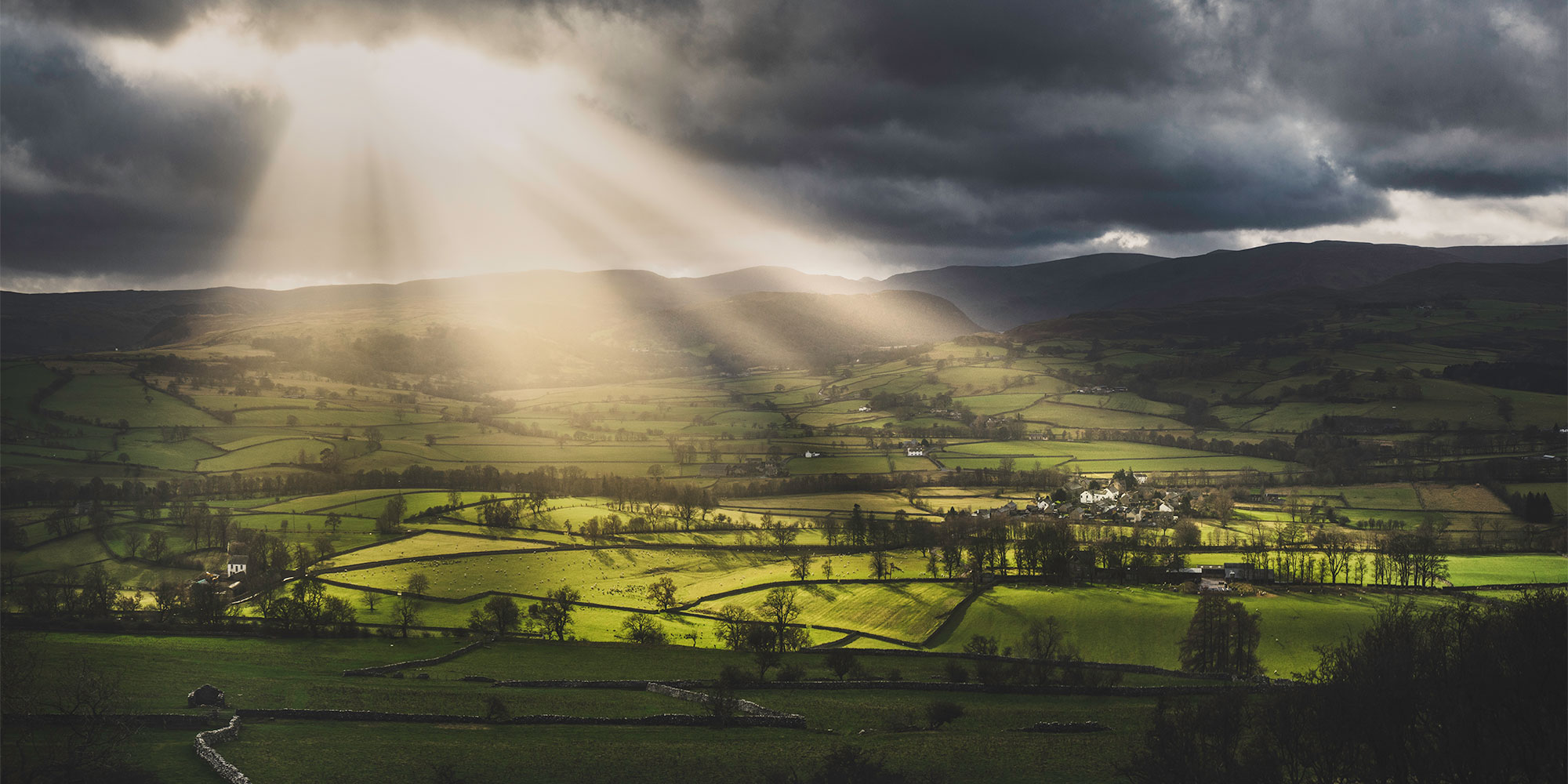 Aerial view of a lush green valley with sunlight piercing through dark clouds, showcasing a serene landscape.