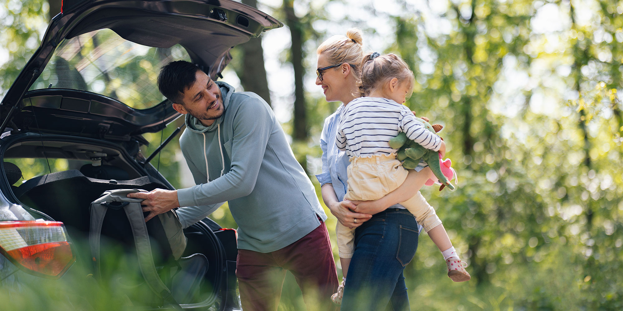 A man packs a backpack into an open car trunk while a woman nearby holds a young child carrying stuffed animals outdoors.