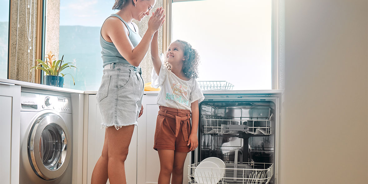 Woman and young girl high-fiving in front of an open dishwasher while it's sunny outside.