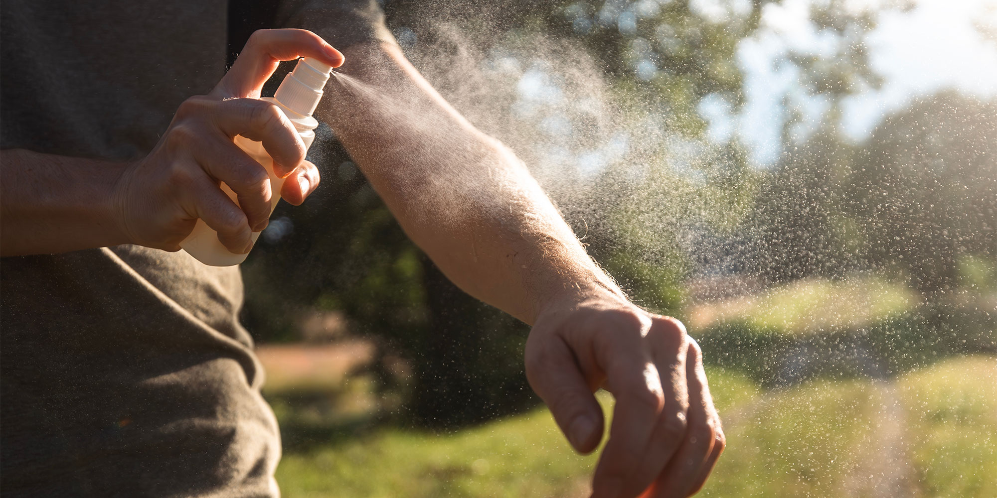A man spraying mosquito repellent onto his arm on a bright day in the countryside.