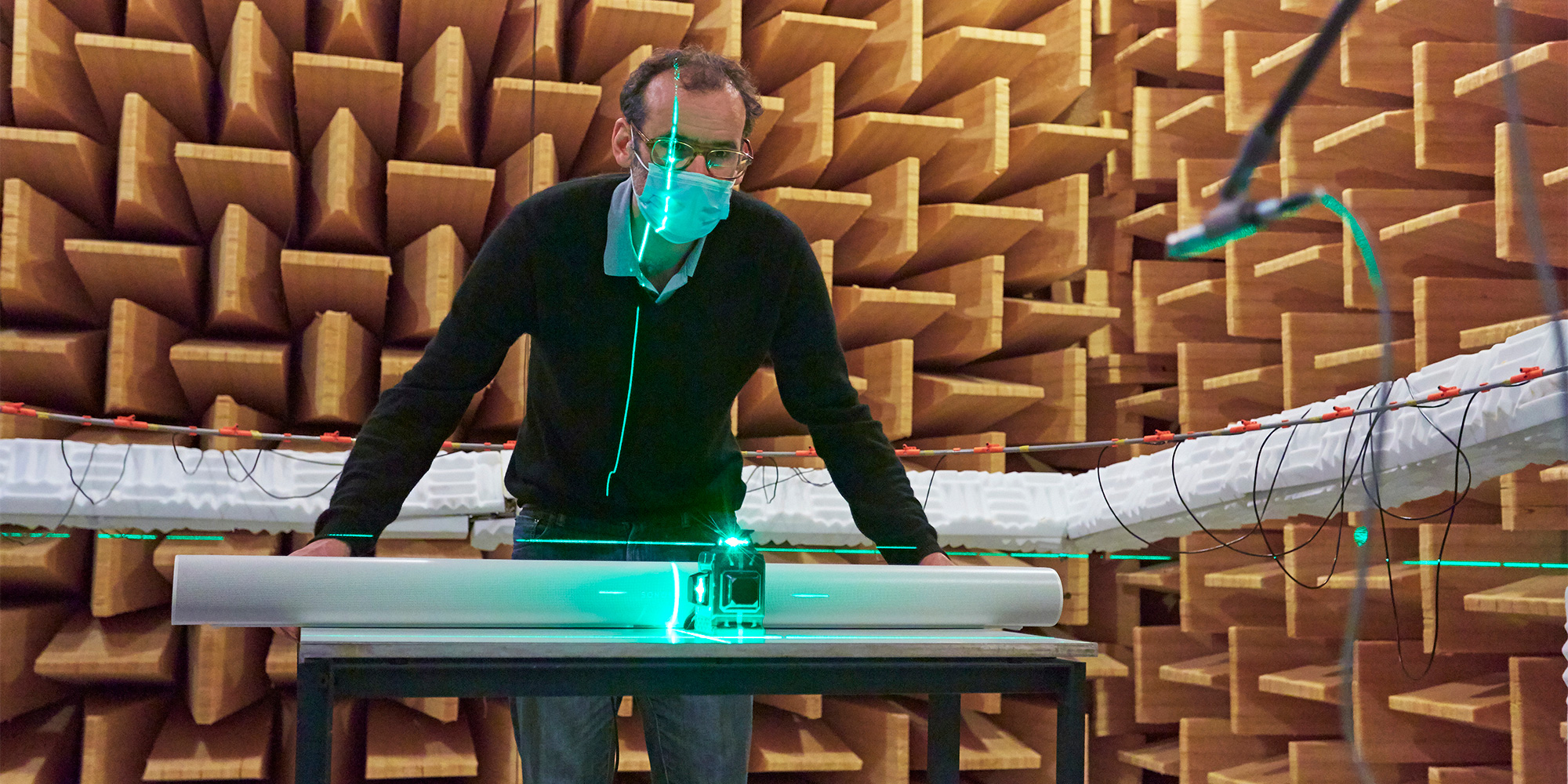 Man stands in front of table lining up a soundbar using a green laser light, in a room with wooden acoustic panels.