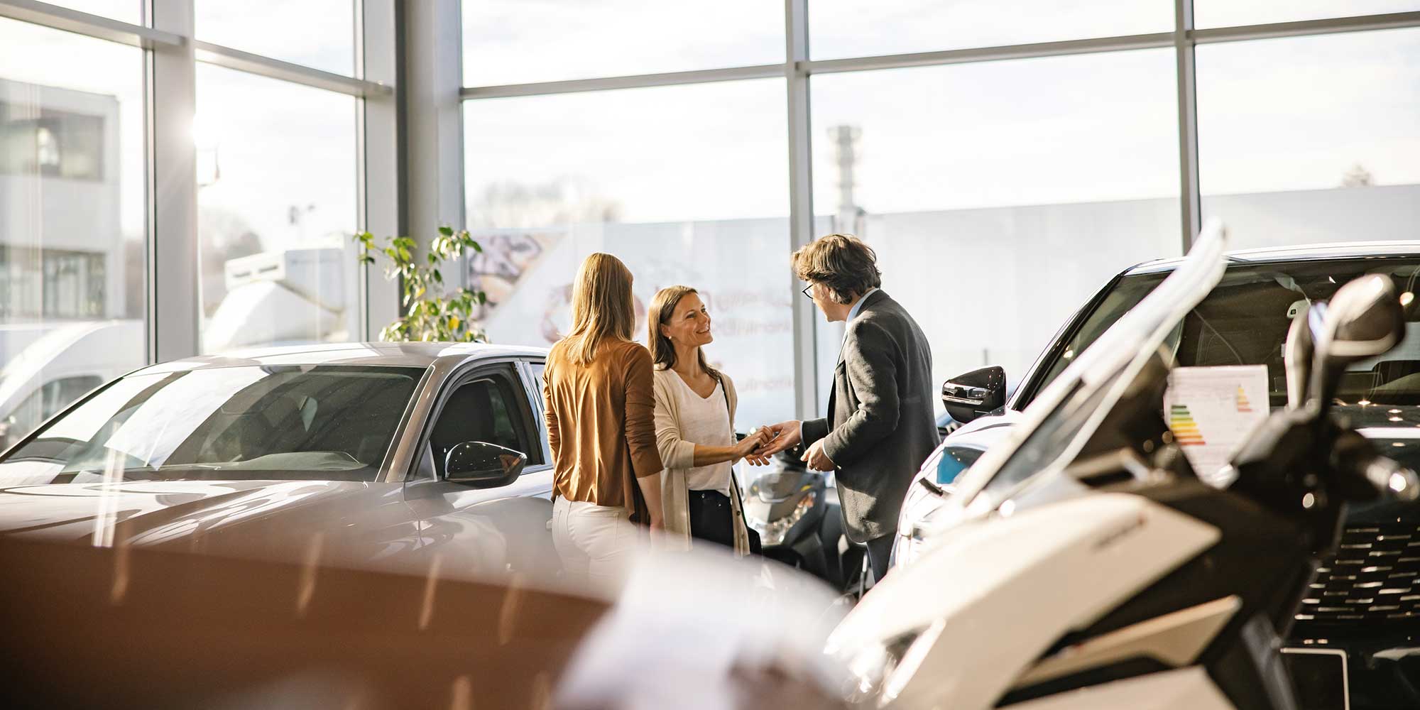 Two women and a man shake hands in a car showroom with several vehicles.