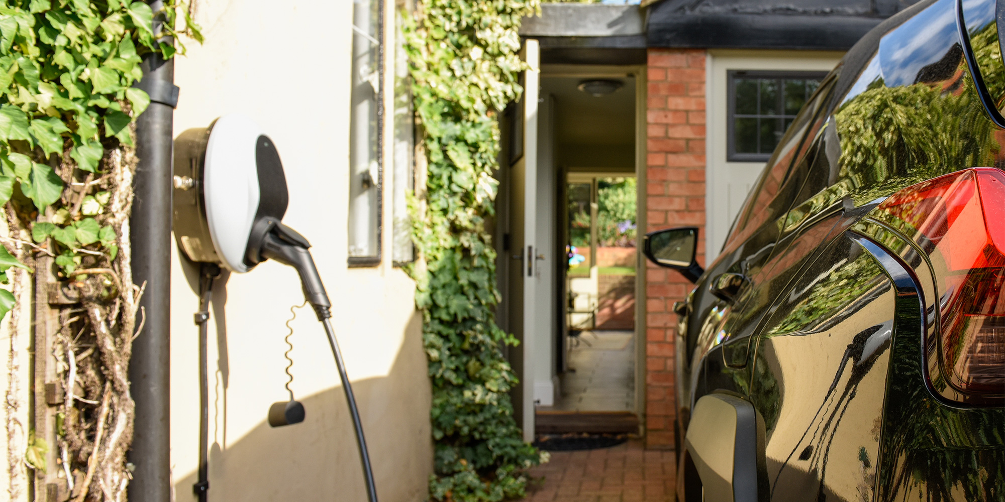 Black car parked at a charging station outside a house with a brick wall and greenery.