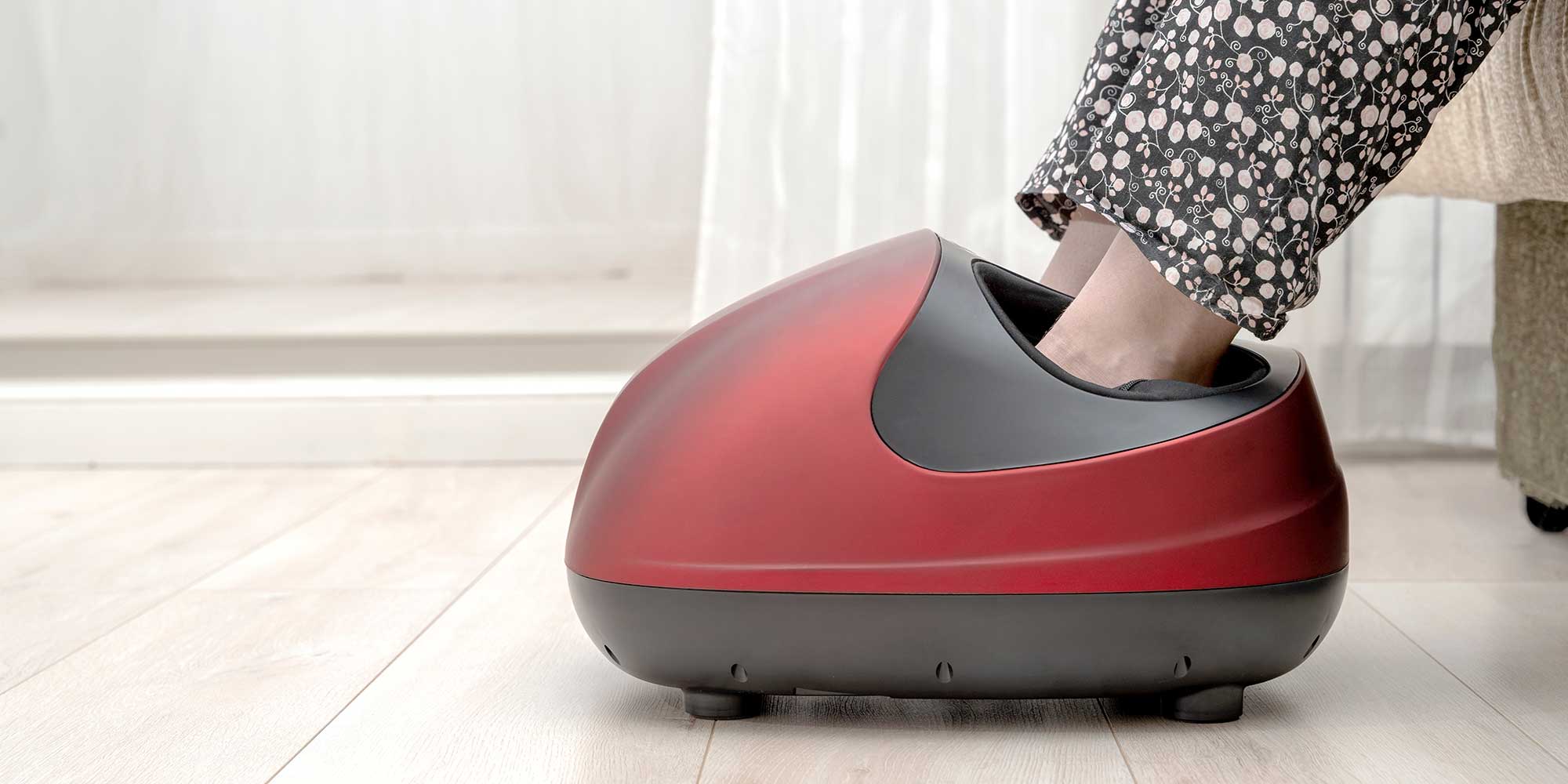 Person in black and white floral pants using a red and black foot massager on a wooden floor.