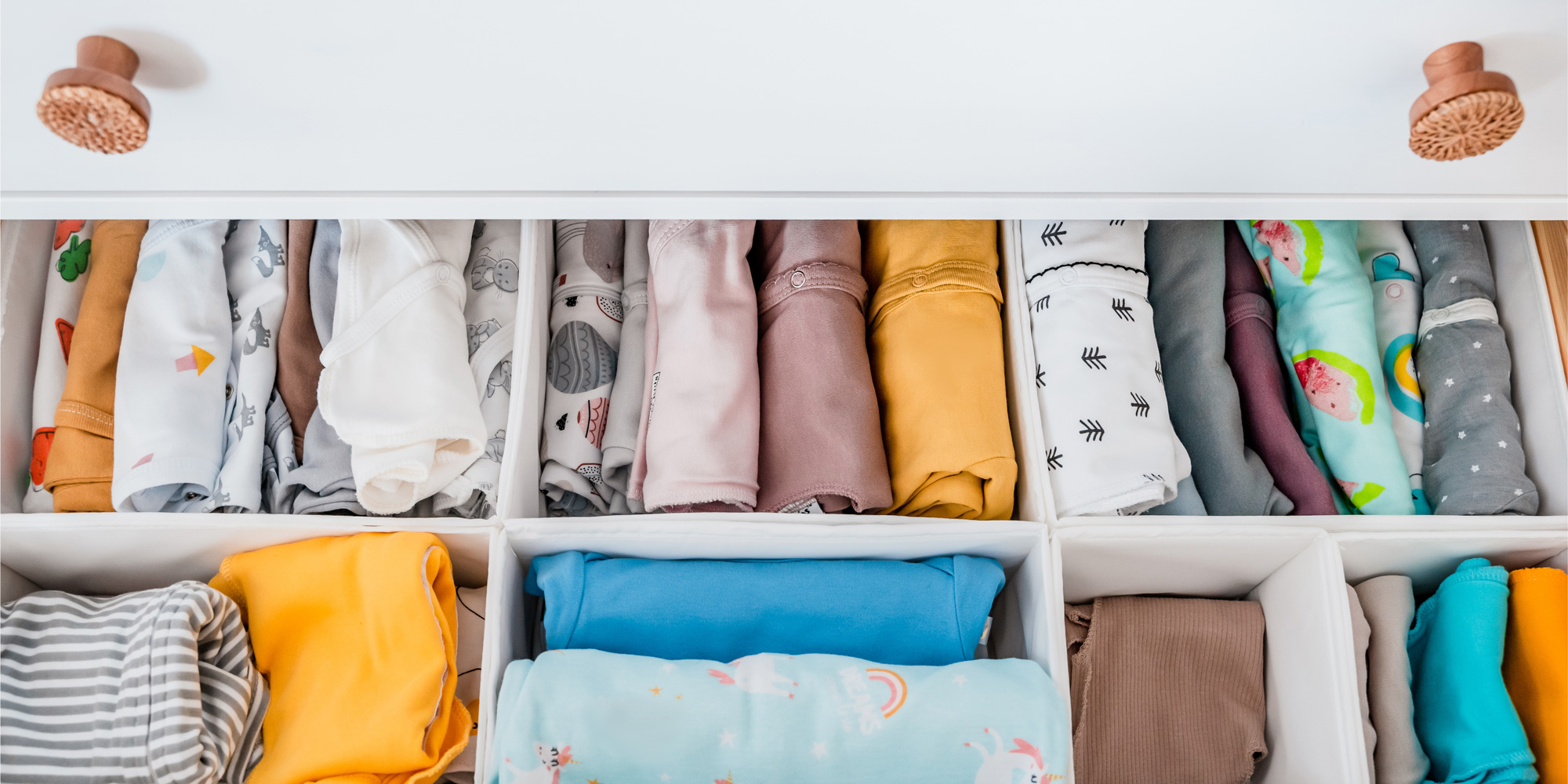A top-down view of an open white dresser drawer neatly organised with various colourful and patterned baby onesies and clothes.