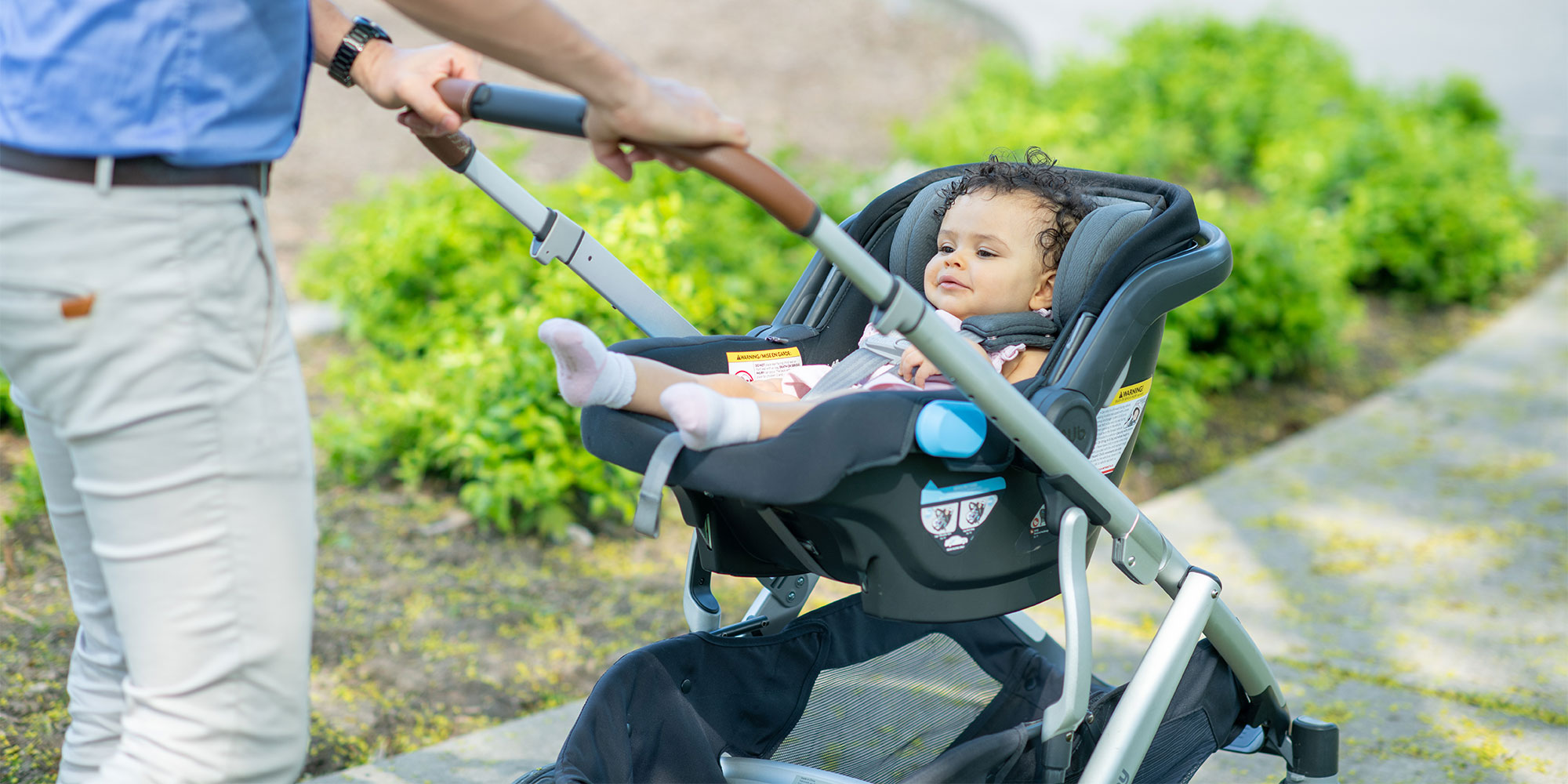 A close-up of a baby sitting in an infant car seat that is docked onto a stroller frame. A person's hands are visible on the brown leather-wrapped handlebar.
