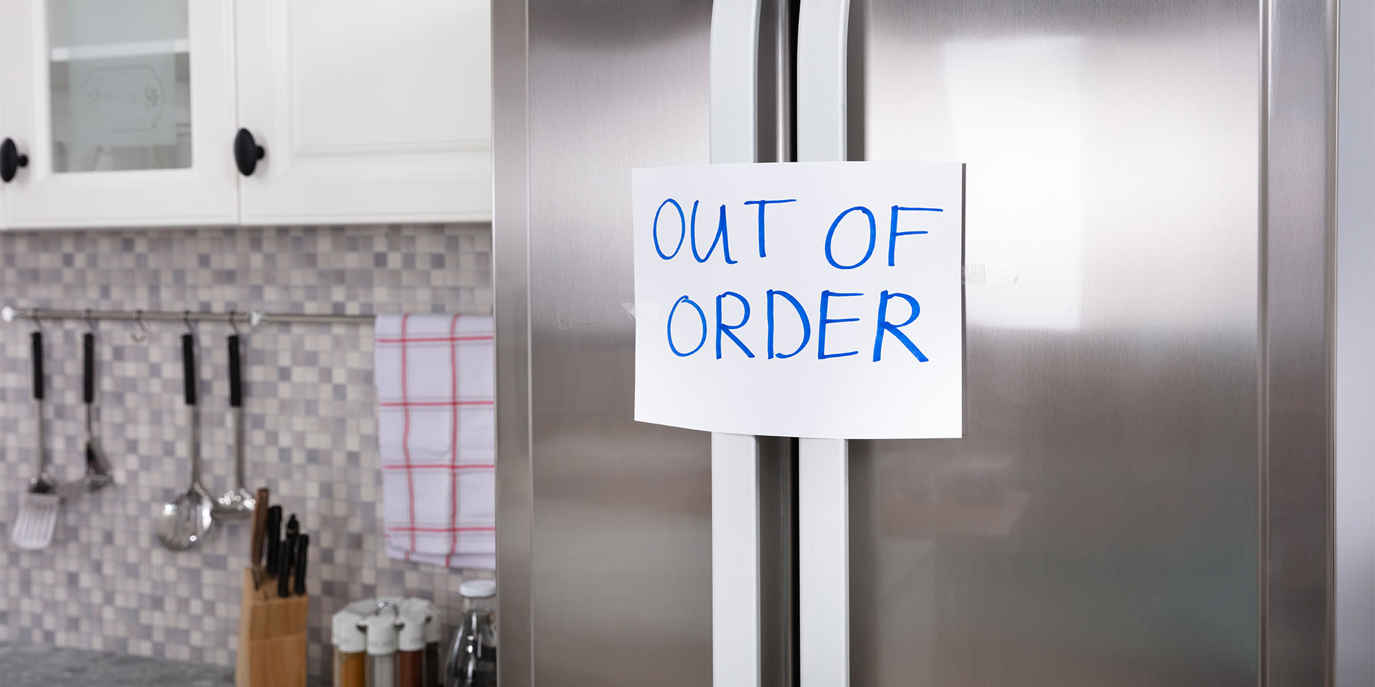 A handwritten 'OUT OF ORDER' sign"is taped across the handles of a closed stainless steel, French-door fridge freezer in a kitchen.