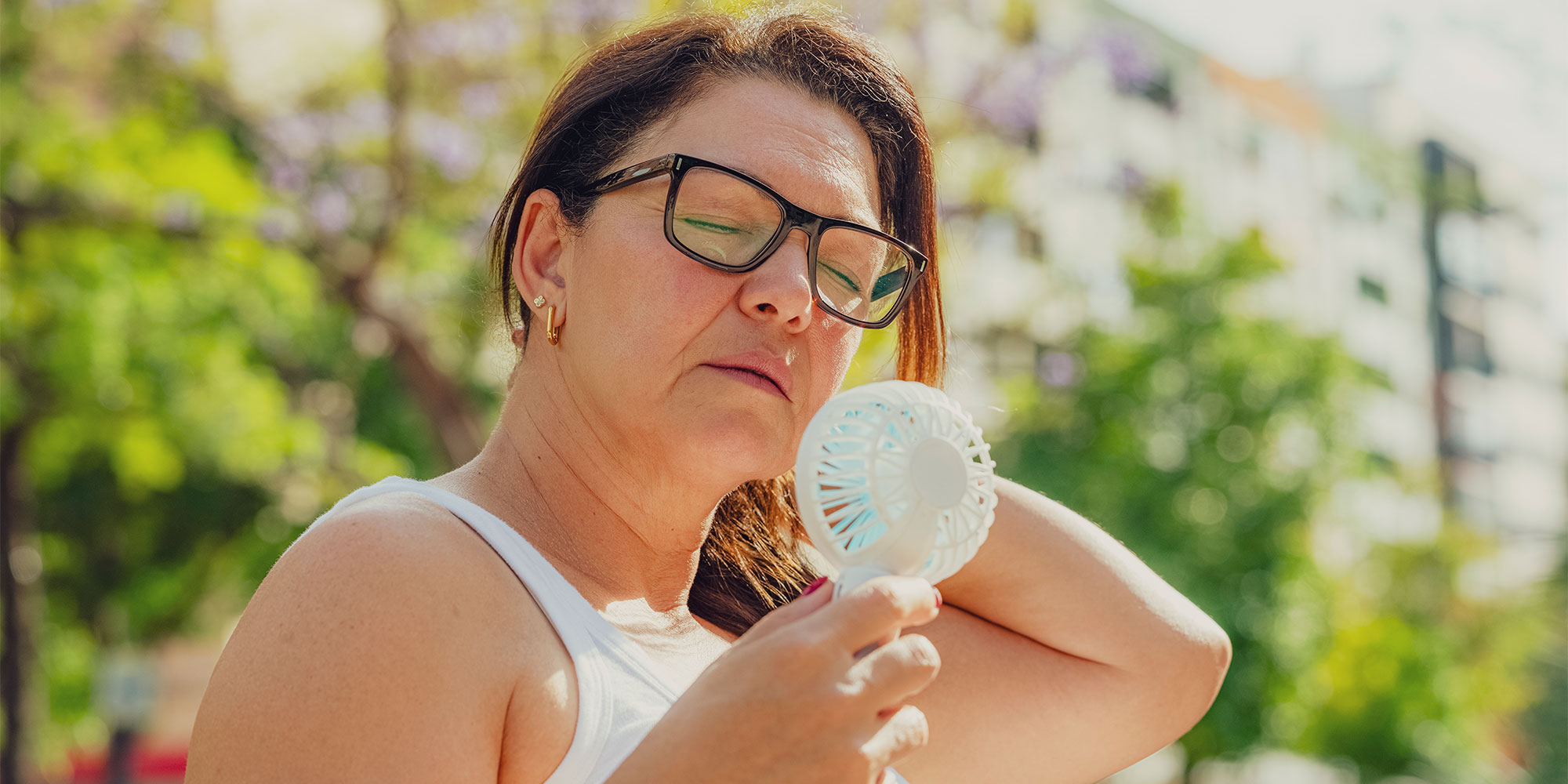 Woman in white tank top and glasses using a portable fan outdoors.