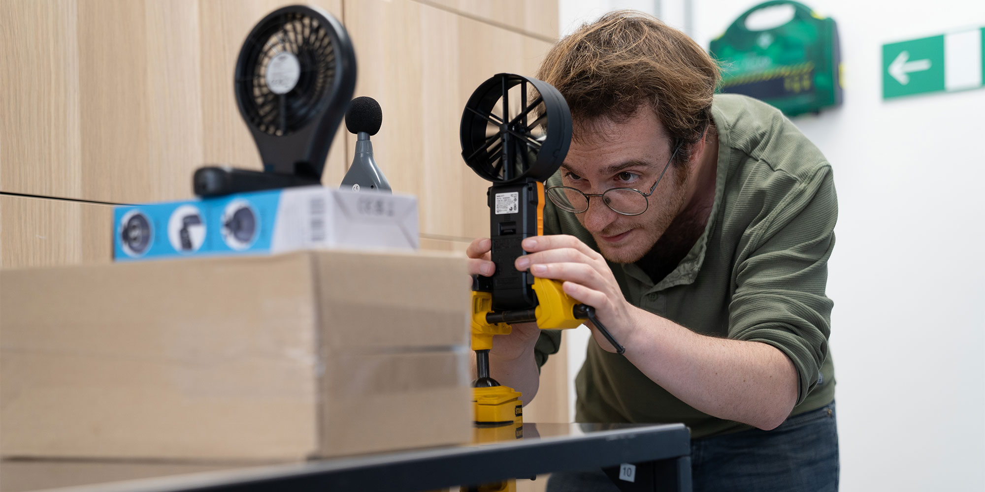 Man in green shirt and glasses using a handheld anemometer to measure wind speed of a handheld fan.