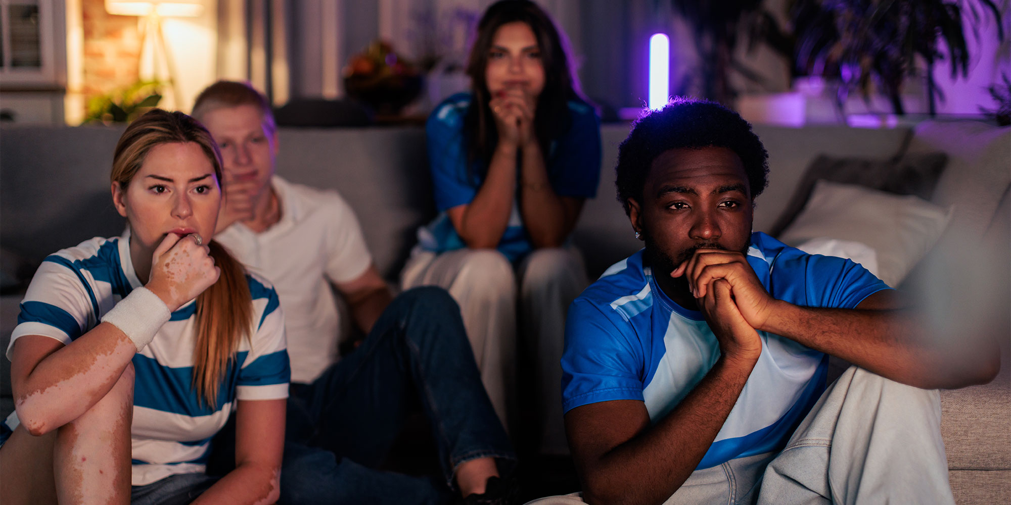 Two men and two women wearing football shirts are sat in front of a TV looking nervous and excited