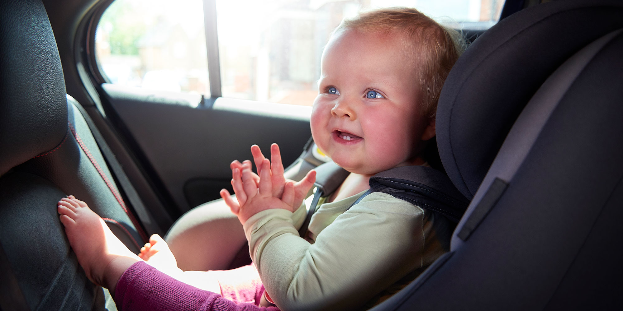 A happy toddler with bright blue eyes smiling and clapping their hands while strapped into a car seat.