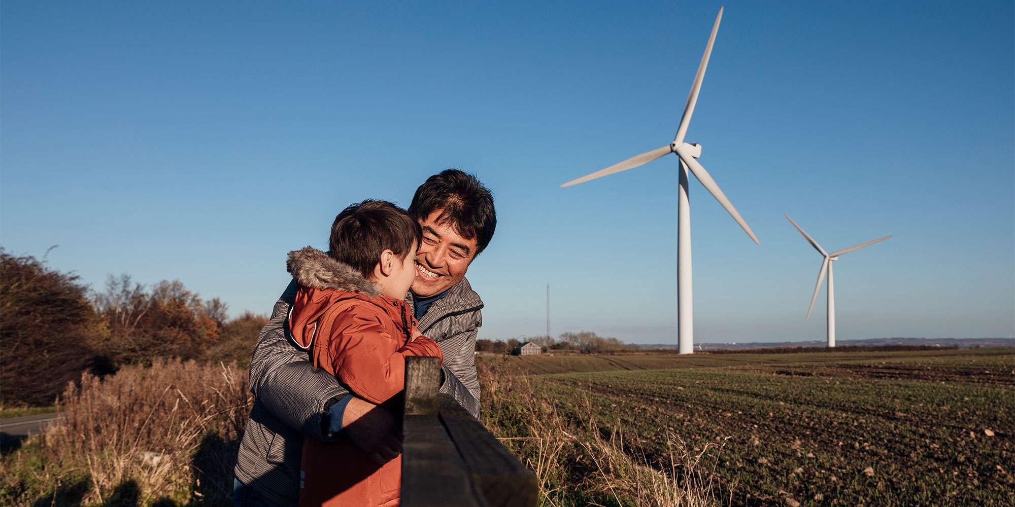 Man and child hugging by a fence in front of wind turbines generating renewable electricity.