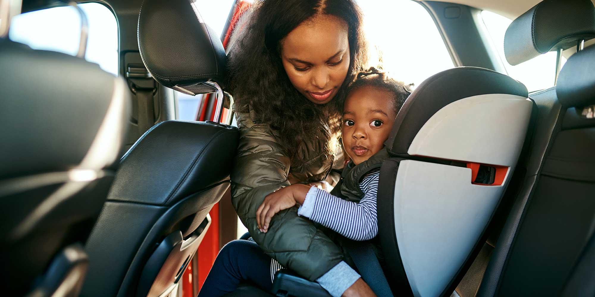 A mother leaning into the backseat of a car, adjusting or securing her toddler in a safety car seat.
