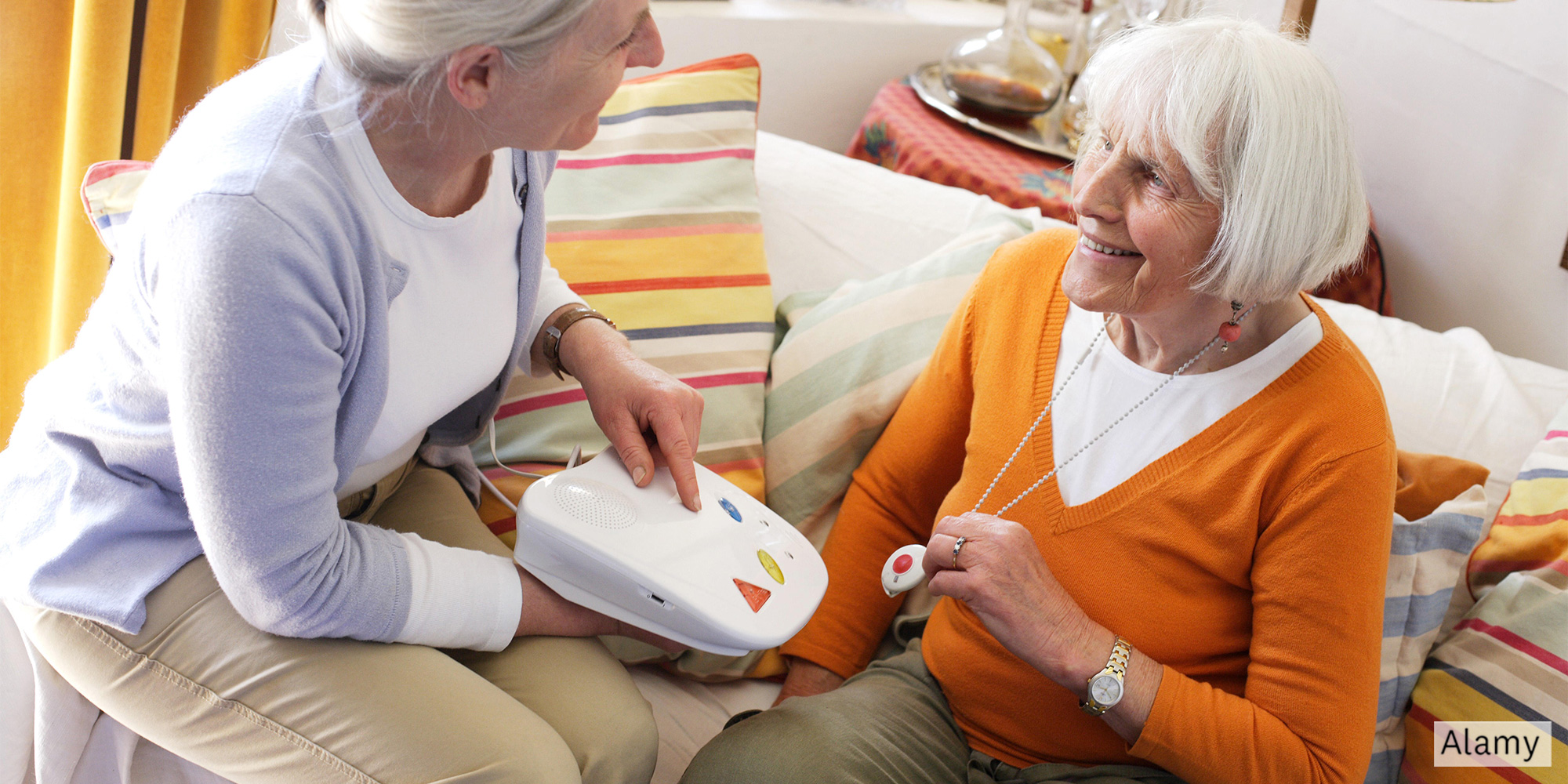 One woman holding a pendant personal alarm and another woman holding the alarm's base unit, both sitting on a sofa