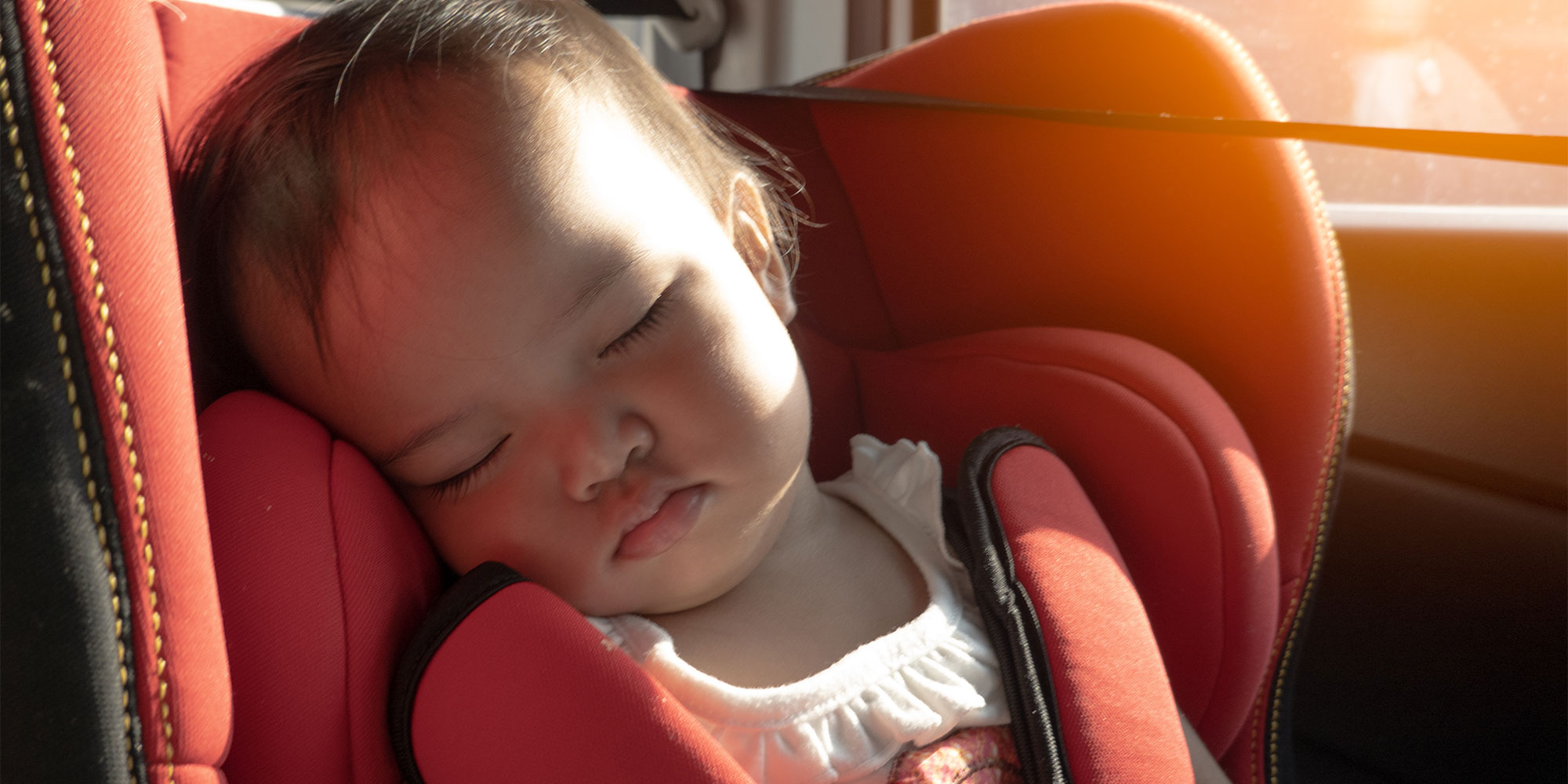 Close-up of a baby sleeping peacefully while buckled into a red and black car seat near a bright window.