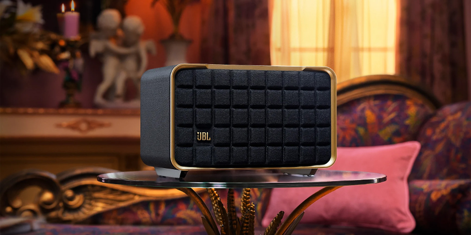 Black JBL speaker with gold accents on glass table in front of ornate couch with pink pillow.