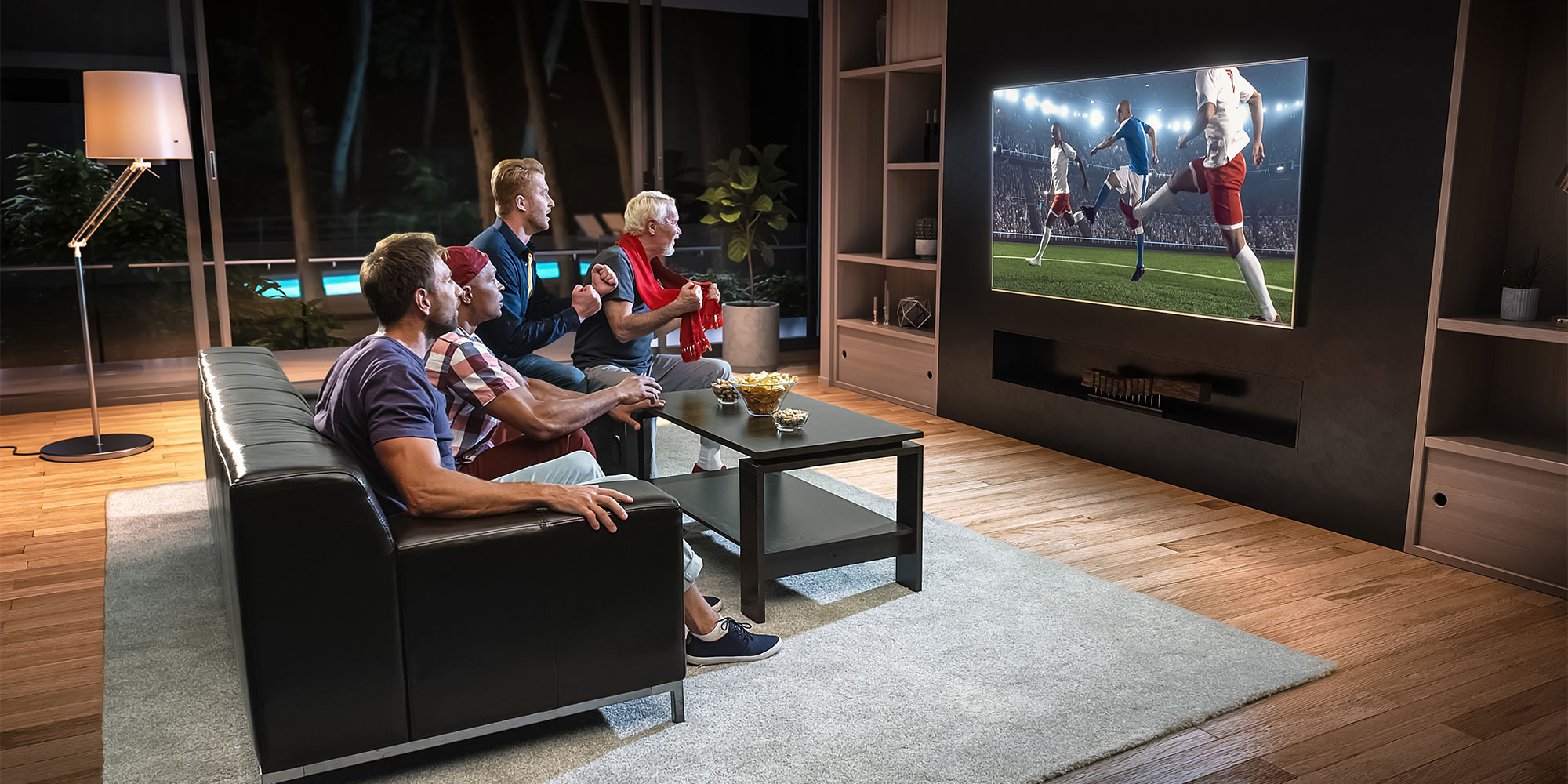 Four men are watching a football match on a large, wall-mounted TV. They appear excited as if their team is about to score.