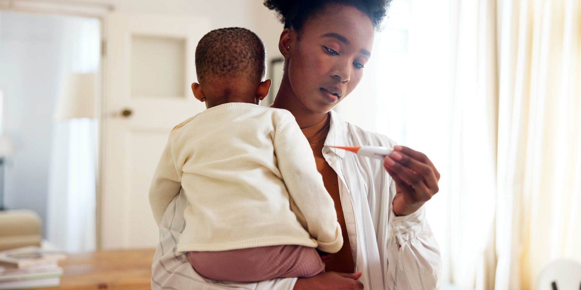 A woman holds a young child in her arms while looking down at a digital thermometer in her hand with a concerned expression.