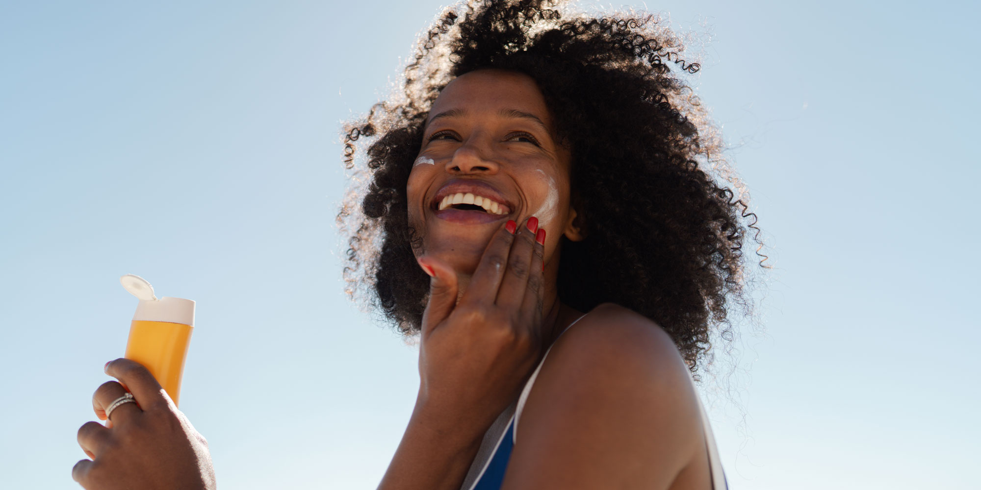 A smiling Black woman with voluminous, curly hair applying sunscreen to her face against a bright blue sky, holding an orange bottle
